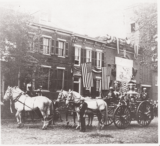 Historic photograph depicting a horse-drawn steam engine on the streets of Alexandria, Virginia. These steam engines were first introduced to the city during the American Civil War, brought in by Union troops trained as firefighters before the war. Before the arrival of steam engines, Alexandria's fire apparatus was pulled to fire by up to twenty-two volunteer firefighters. This image illustrates the evolution of firefighting technology and the transition from human-powered to mechanized equipment in Alexandria during the 19th century.