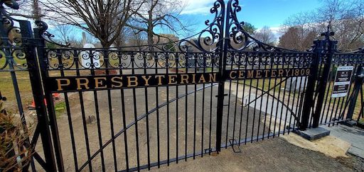 Main entrance gate of Presbyterian Cemetery in Alexandria, Virginia. The black wrought iron Victorian-style gate, built in 1903, features ornate scrollwork and gold lettering reading “Presbyterian Cemetery 1809.” Trees and gravestones are visible inside the fenced grounds beyond the gate.