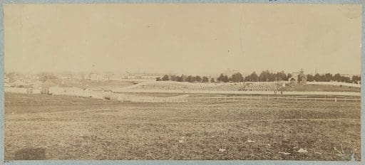 Historic 1865 photograph of Alexandria National Cemetery taken from West Village across Hooff’s Run. The cemetery’s entrance arch is visible in the distance, though its inscription cannot be read from this angle. A large open field dominates the foreground, reflecting the undeveloped landscape surrounding the cemetery at the time. The vantage point is likely near the 1800 block of Jamieson Avenue, along the former route of the Orange & Alexandria Railroad. Image courtesy of the Library of Congress.