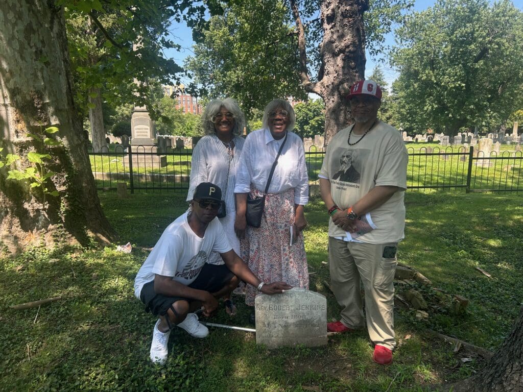 Dr. Darnella McGuire Nelson with her mother, husband, and a family member beside the gravestone of Reverend Robert Jenkins in Douglass Cemetery, part of the Wilkes Street Cemetery Complex.