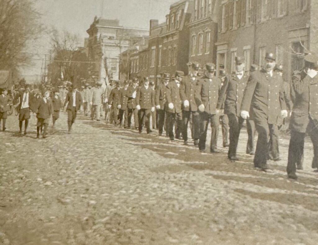 Funeral procession for Fire Chief George W. Petty with a flower-covered casket on a historic hose reel drawn by 50 firemen, passing fire stations on Prince Street as city and firehouse bells toll in tribute."