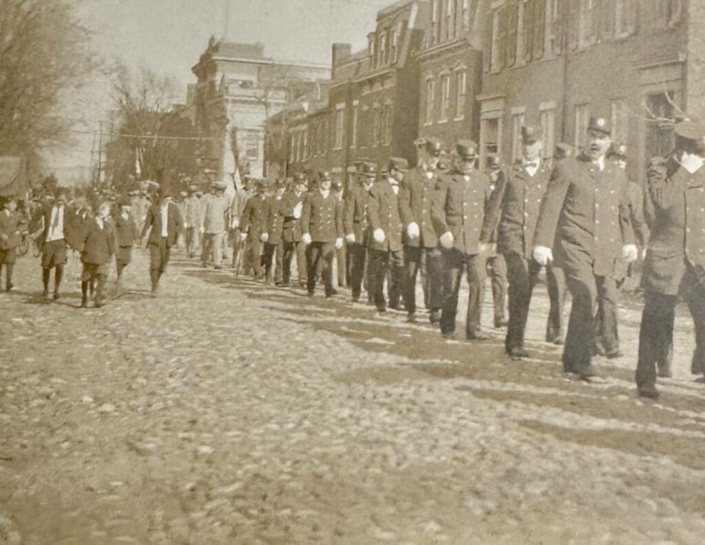 Funeral procession for Fire Chief George W. Petty with a flower-covered casket on a historic hose reel drawn by 50 firemen, passing fire stations on Prince Street as city and firehouse bells toll in tribute."