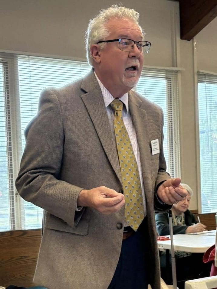 David Heiby, public historian, stands and gestures while giving a presentation. He is wearing a tan blazer, patterned yellow tie, and glasses, speaking to an attentive audience seated at tables in the background.