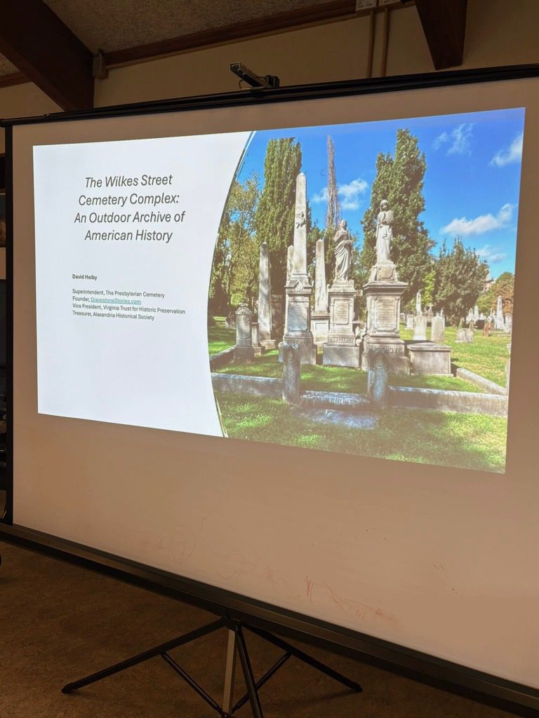 Opening slide of a PowerPoint presentation titled “The Wilkes Street Cemetery Complex: An Outdoor Archive of American History” by historian David Heiby. The slide shows a photograph of tall nineteenth-century monuments and statues in Alexandria’s Wilkes Street Cemetery Complex beneath a bright blue sky.