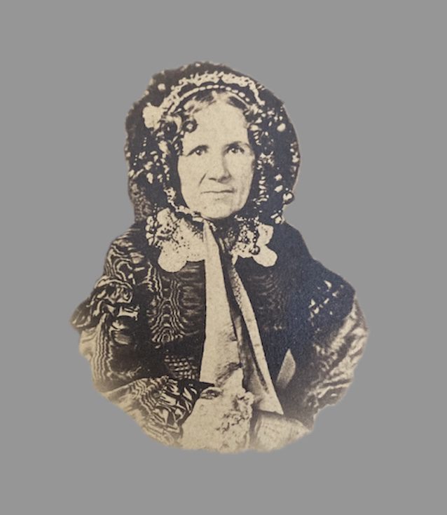 Sepia-toned photograph of Julia Anna Marion Prosser Lee in later life. She is shown seated, wearing a patterned dark dress with long sleeves and a bonnet trimmed with lace and ribbons. Her face is framed by curls beneath the bonnet, and she gazes calmly at the camera.