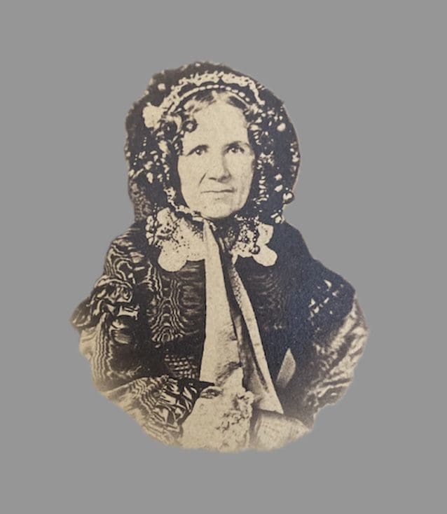 Sepia-toned photograph of Julia Anna Marion Prosser Lee in later life. She is shown seated, wearing a patterned dark dress with long sleeves and a bonnet trimmed with lace and ribbons. Her face is framed by curls beneath the bonnet, and she gazes calmly at the camera.