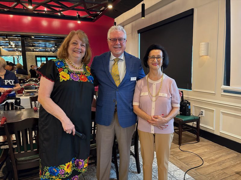 Three people pose together in a restaurant setting - a woman in a colorful floral dress on the left, a man in a blue suit jacket and yellow tie in the center, and a woman in a pink top on the right, all wearing name tags and smiling at the camera.