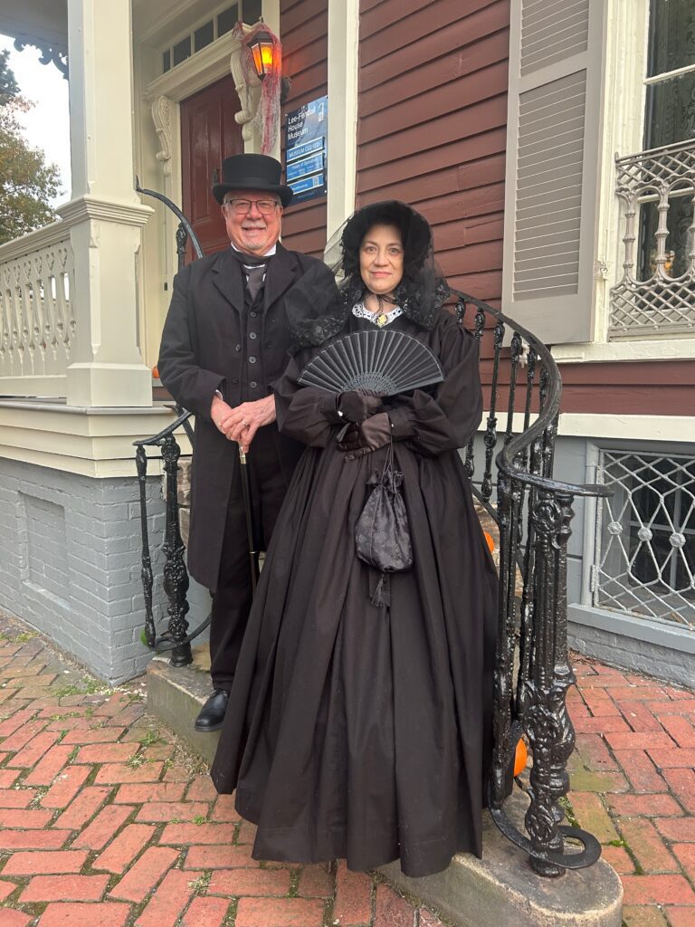 David Heiby and Catherine Weinraub dressed in 19th-century attire on the steps of the Lee-Fendall House Museum in Alexandria, Virginia. David, portraying Mayor E. E. Downham, wears a black frock coat and top hat, while Catherine, in mourning dress as a Victorian widow, holds a black fan and reticule.