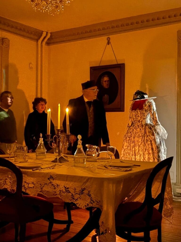 David Heiby portraying E. E. Downham in the candlelit Federal-style dining room of the Lee-Fendall House, with the portrait of Philip Richard Fendall II on the wall and a period dress by Emma Rowland displayed on a dress form nearby.