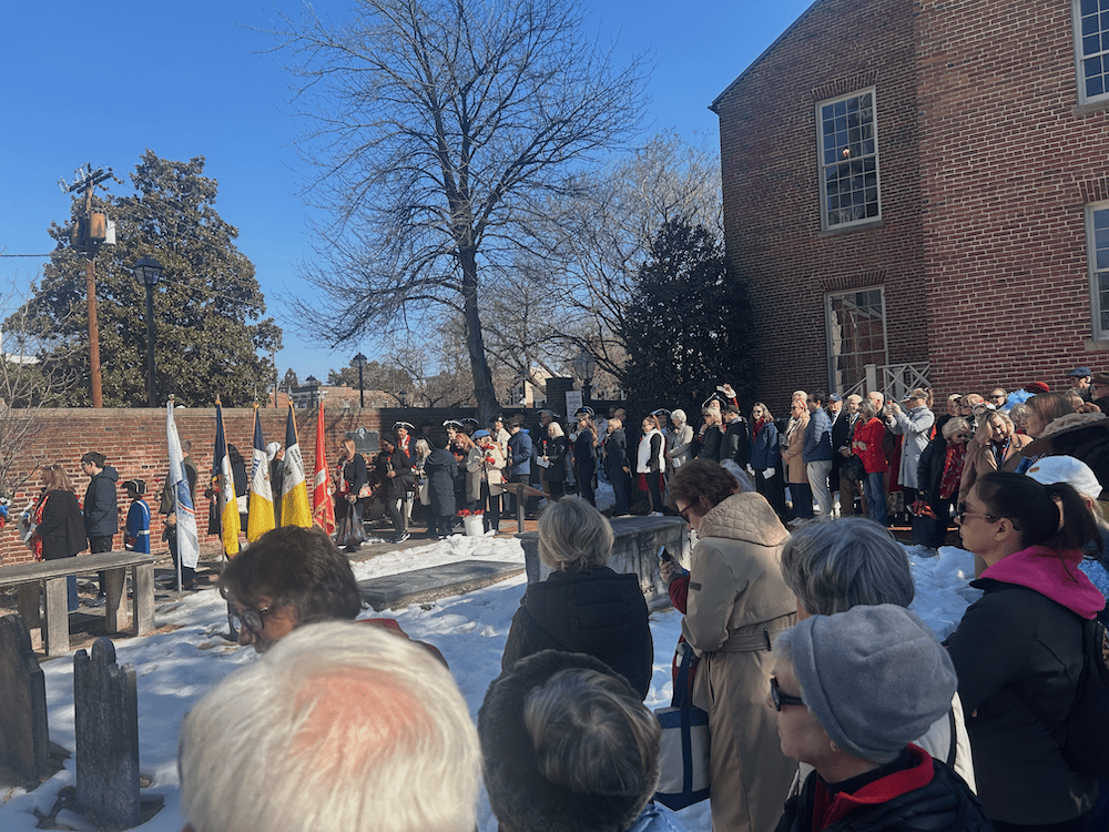 Crowd gathered along cleared sidewalks inside the Old Presbyterian Meeting House burial ground in Alexandria, Virginia, during the February 14, 2026 Wreath Laying Ceremony, with snow covering parts of the cemetery and flags displayed near the tomb.
